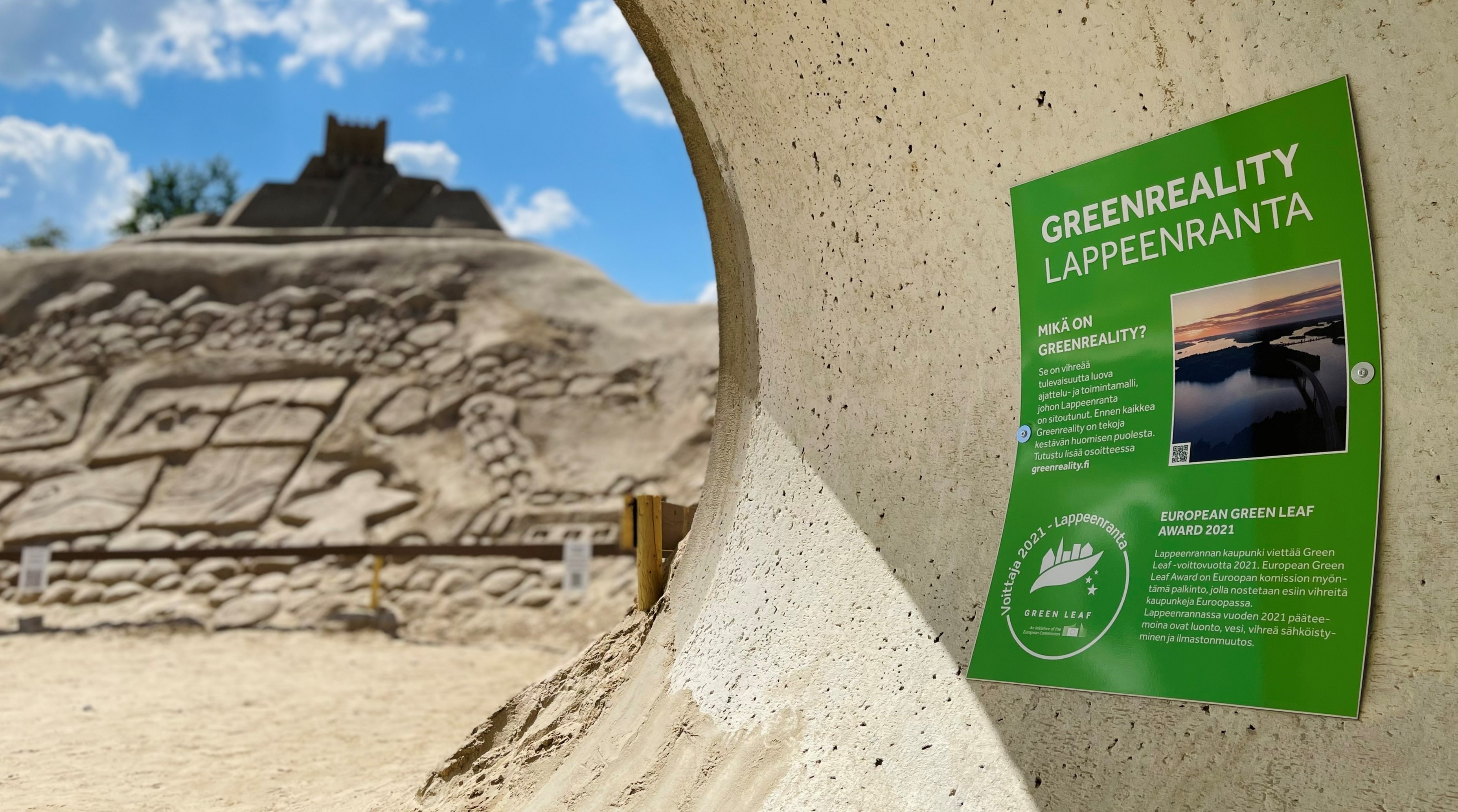 Vihreä infotaulu, jossa lukee suurella tekstillä Greenreality Lappeenranta. Taustalla sumeana hiekkalinnan veistoksia ja sinistä taivasta.