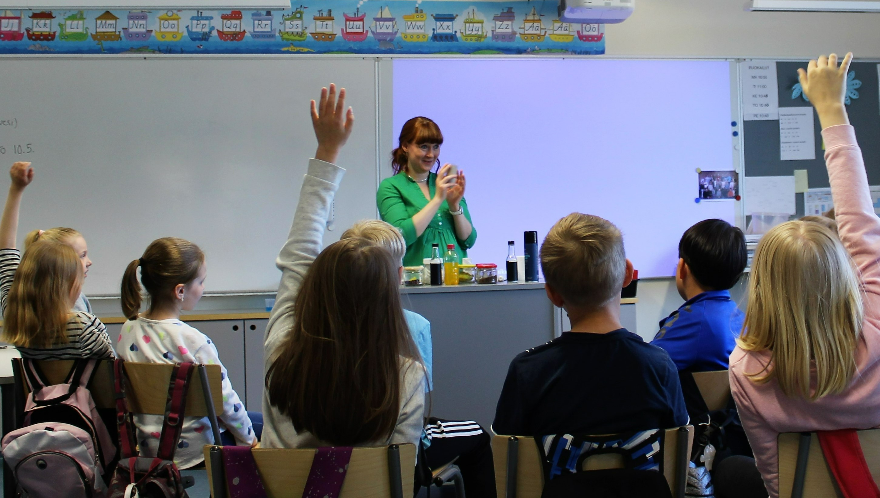 Students in a classroom. The teacher is in the front of the room, smiling in the direction of the students.