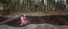 Pump track cycle track, with a man riding a mountain bike and a little girl biking in a pink outfit with fairy wings on her back. Forest in the background.