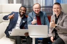 Three smiling men sitting with laptops in front of them, showing thumbs up.