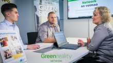 Kaksi miestä ja nainen istuvat neuvottelupöydän ääressä. Taustalla Greenreality Networkin markkinointimateriaaleja. Alareunassa vihreä Greenreality Network -logo.