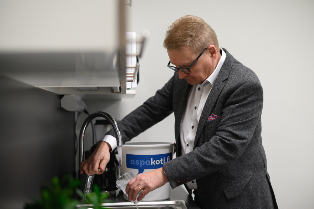 A man pours water from a curved faucet into a drinking glass. In the back, a bucket with the text aspakoti.fi.