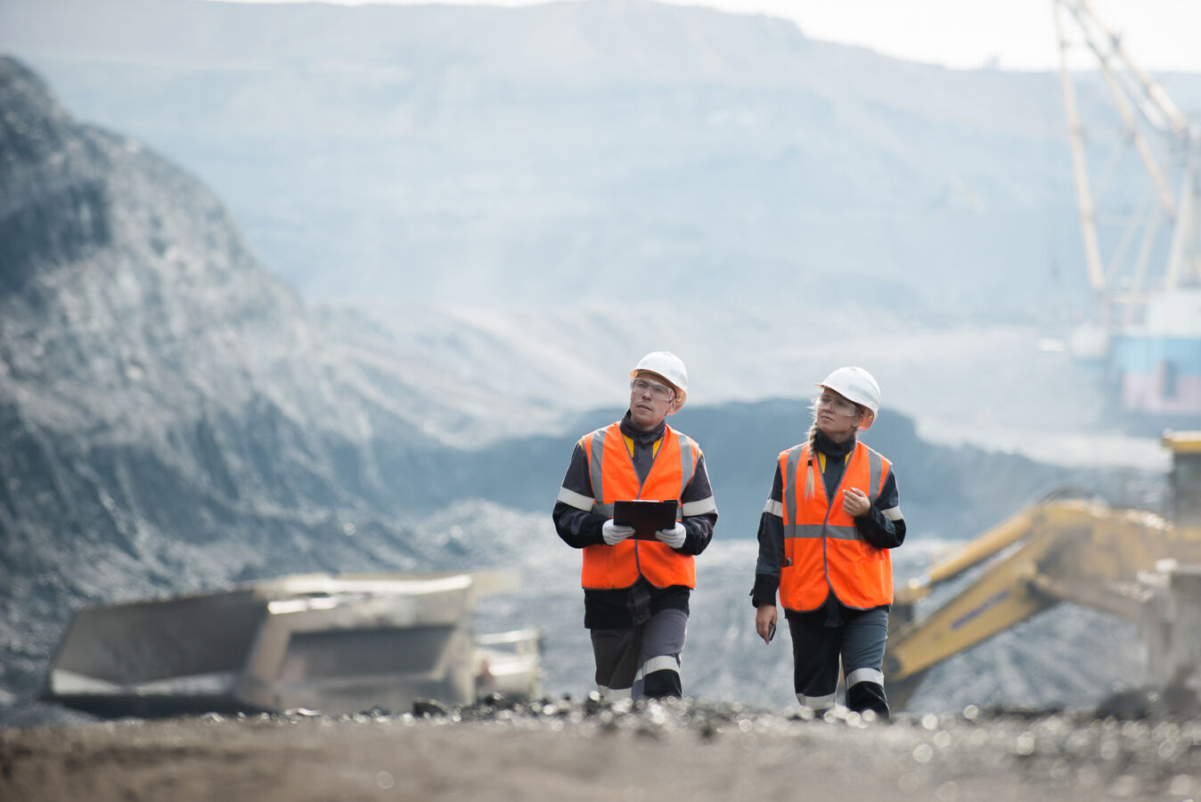 A quarry, working machines, a man and a woman wearing helmets and safety vests walking side by side.