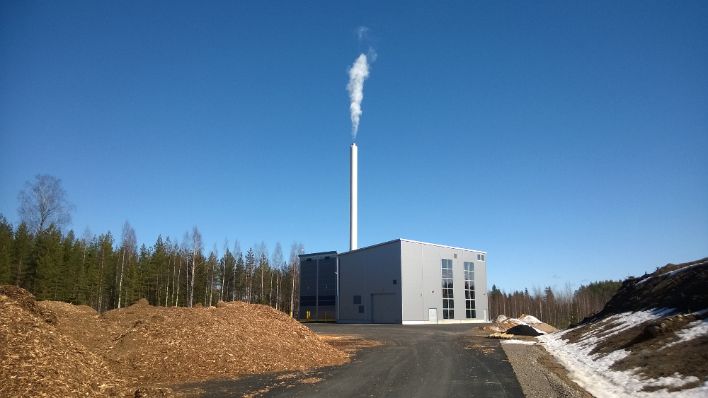 Energialaitos, jonka piipusta tulee savua. Etualalla hakekasa. Taustalla metsää ja sininen taivas.